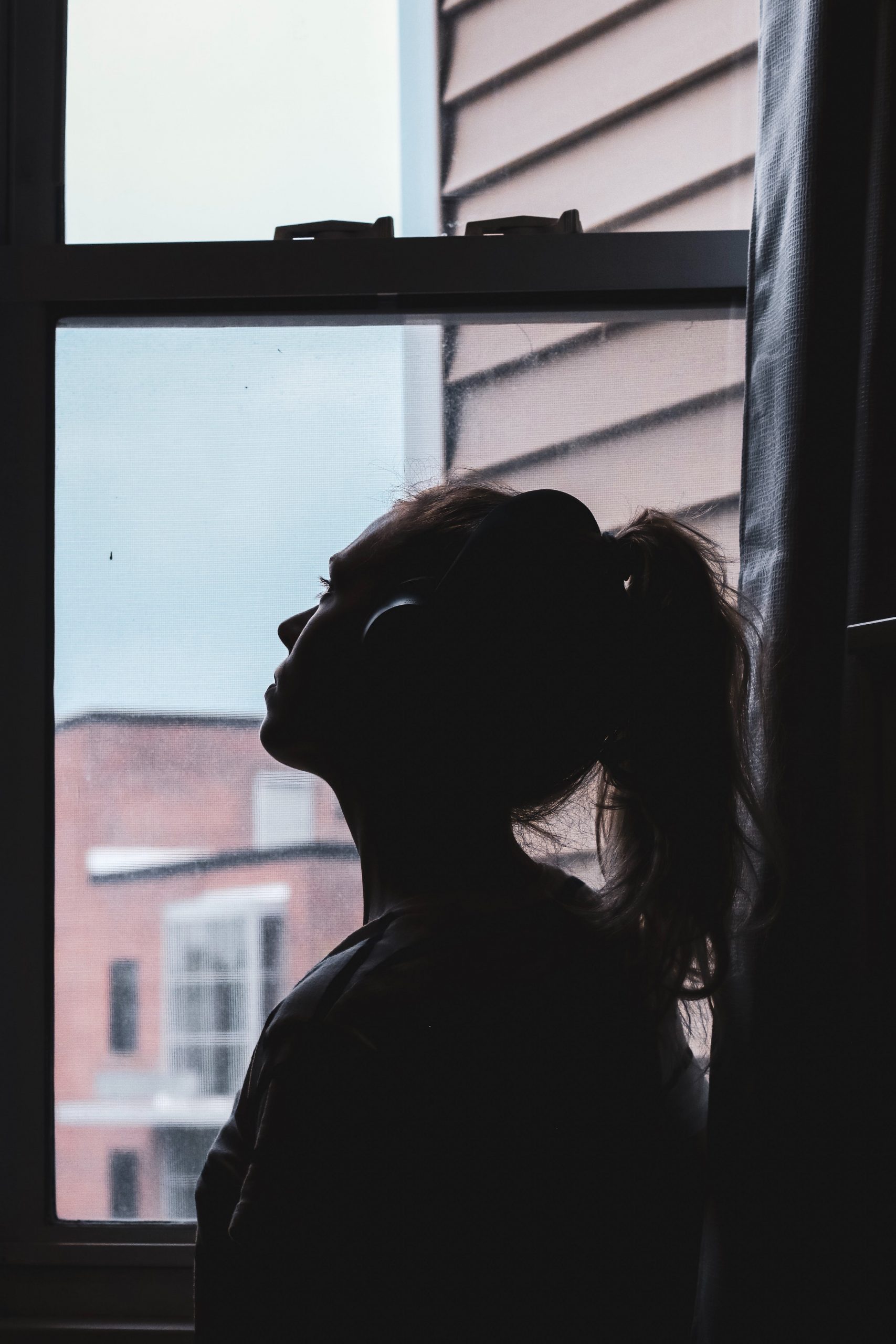 A woman with headphones is looking at the gloomy weather through the window .