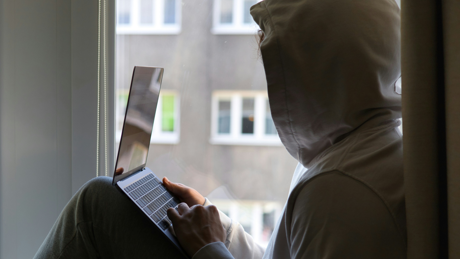 Side view of a person in a light neutral hoodie sitting in front of a laptop by a window. Their face is fully covered by the hood, symbolizing potential isolation in the digital age.