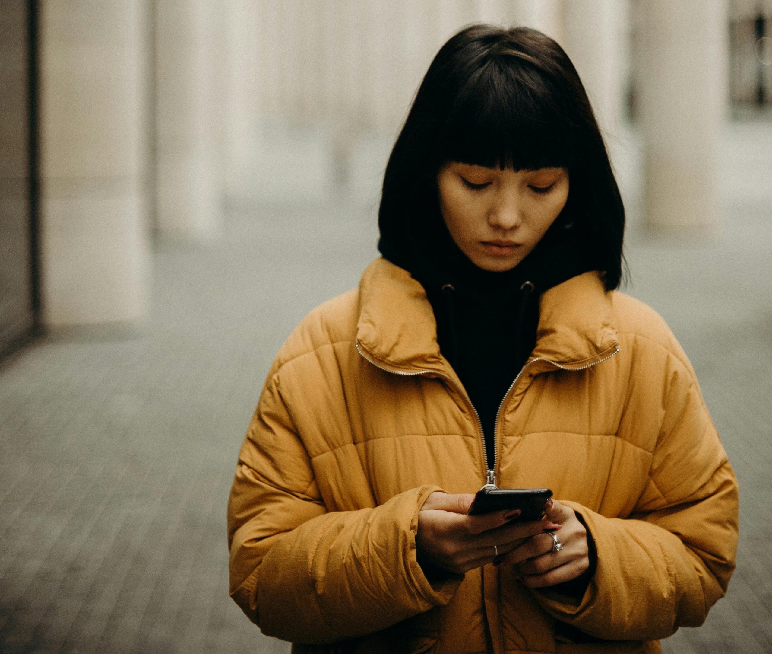 Sad woman in a yellow jacket looking at the screen of her phone.